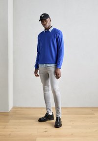 Suéter de manga larga azul sobre una camisa de cuadros azul claro, jeans grises ajustados y zapatos negros. Gorra negra con un logo blanco en la parte delantera.