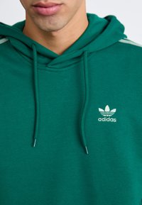 Grønn bomullslue med snor, med en liten hvit Adidas-logo på brystet og teksturert stoff.