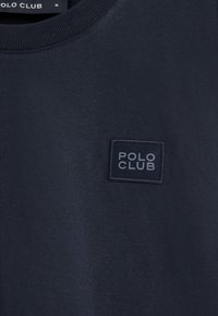 Marineblauer Pullover mit geripptem Halsausschnitt und einem rechteckigen Stoffpatch, auf dem der Text "POLO CLUB" in einer helleren Nuance steht. Weiche Textur.