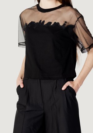 Woman wearing a black sheer short-sleeve top with "womani" embroidery and black pleated trousers, hands in pockets.