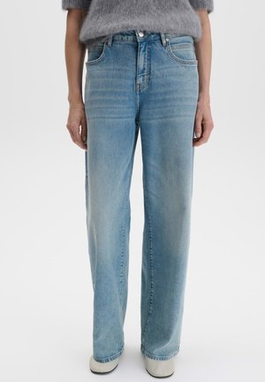 Person trägt hellblaue Jeans mit weitem Bein und weiße Schuhe, kombiniert mit einem grauen, flauschigen Kurzarmtop, steht vor einem einfarbigen Hintergrund.