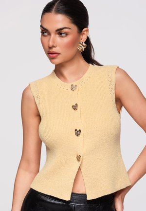 Kobieta w żółtym, bezrękawnym sweterku z guzikami w kształcie serc i dużymi złotymi kolczykami, pozująca z jedną ręką na biodrze.