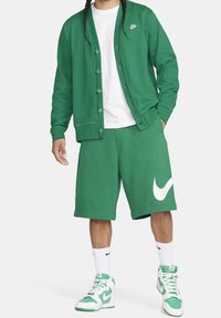 Osoba ve zeleném knoflíkovém svetru Nike, ladících zelených šortkách s velkým bílým swooshem, bílém tričku, bílých ponožkách Nike a zeleno-bílých teniskách.