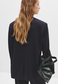 Kvinna med lång röd hår som bär en mörk rutig blazer och håller en svart läderhandväska, vänd på sidan mot en enkel vit bakgrund.