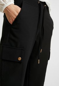 Mustat cargo-housut, jotka on valmistettu teksturoidusta kankais- ta, ja joissa on sivutasku kultanapilla ja säädettävät narut vyötäröllä.