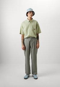 Lichtgroen kortarm overhemd met knopen en een gestructureerd patroon, grijze broek en blauwe bucket hat. Tan schoenen met blauwe accenten.