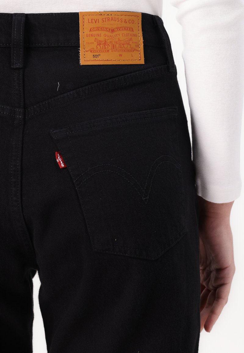 Jeans de mezclilla negra con un parche de cuero que presenta texto en relieve. Las características notables incluyen una etiqueta roja en el bolsillo y un diseño de costura sutil.