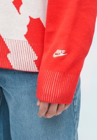 Crvena pletena džemper s bijelim apstraktnim uzorcima, rebrastim rukavima s crvenim i bijelim prugama, i malim bijelim Nike logom na rukavu.