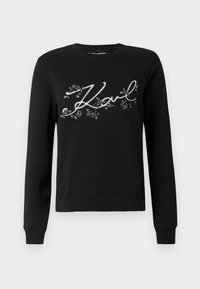 Sudadera negra de manga larga y cuello redondo, con acentos florales y un logo plateado en cursiva en el pecho. Tejido suave y texturizado.