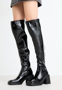 Bottes noires en cuir verni montant jusqu'aux genoux, à talons blocs, dotées d'une fermeture éclair latérale, d'une texture lisse et d'un design élégant et ajusté.