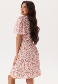 Blommig klänning med korta puffärmar, tillverkad av lättviktsmaterial. Har en vit botten med röda blommor och resårmidja för en bra passform.