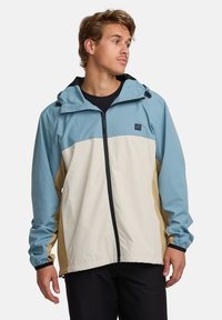 Lightweight, color-blocked jacket in blue, beige, and tan, with a zip front, adjustable hood, and elastic cuffs. Smooth texture, casual design.