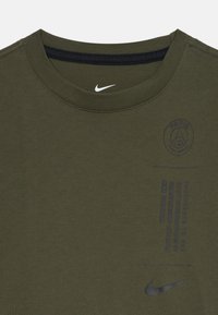 Camiseta Nike verde oliva con cuello redondo, que presenta un logo de París en azul oscuro y texto impreso en un diseño minimalista. Textura de tela suave.