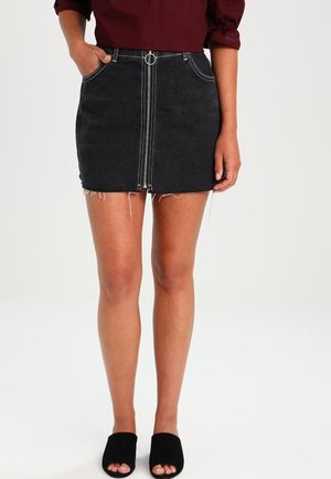 Mini falda de denim negra con cremallera frontal, dobladillo deshilachado, dos bolsillos laterales y detalle de costura blanca, combinada con mule negras de punta abierta.