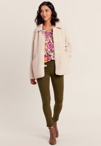 Giacca in pile beige con chiusure a toggle, indossata sopra una blusa floreale. I pantaloni fitted verde oliva e gli stivali alla caviglia beige completano l'outfit.