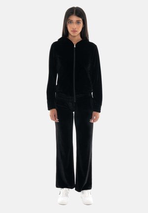 Donna che indossa una felpa nera in velluto con zip e pantaloni a gamba larga abbinati, con sneakers bianche, in piedi di fronte a uno sfondo semplice.