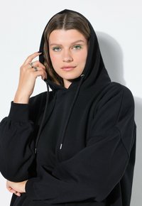 Černá mikina s kapucí na šňůrku, velkými nabíranými rukávy a měkkou texturou. Modelka pózuje s jednou rukou na kapuci.