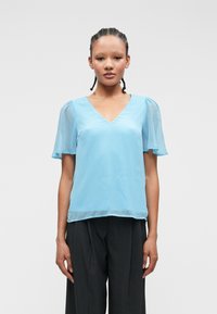 Ανοιχτό μπλε μπλούζα με V λαιμόκοψη και διάφανα κοντά μανίκια. Μαλακό, ρέον ύφασμα με λεία υφή, συνδυασμένο με σκούρες γκρι παντελόνες.