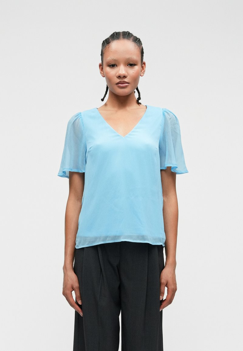 Ανοιχτό μπλε μπλούζα με V λαιμόκοψη και διάφανα κοντά μανίκια. Μαλακό, ρέον ύφασμα με λεία υφή, συνδυασμένο με σκούρες γκρι παντελόνες.