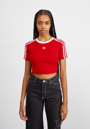 Camiseta corta roja con cuello blanco y logo de Adidas, con diseño de tres rayas blancas en las mangas. Combinada con pantalones negros.