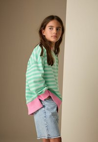 Zelena prugasta majica s ružičastim manžetama, uparena s traper kratkim hlačama. Tkanina izgleda mekano, a dizajn je casual stila.