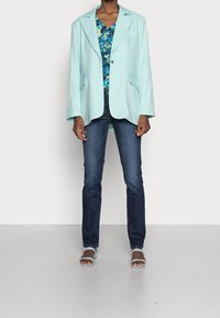 Helles blaues, übergroßes Blazer mit Taschen, getragen über einem blumigen Top. Kombiniert mit dunklen Jeans und blauen Sandalen. Weicher Stoff und lockere Passform.