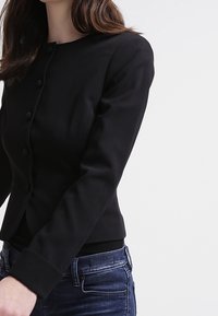 Svart skräddarsydd kavaj med rund halsringning, enfärgad design och textilstruktur; har fem knappar och något vadderade axlar.