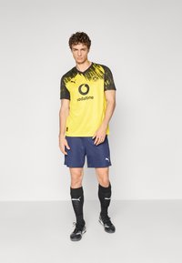 Gul og sort fodboldtrøje med et grafisk mønster, parret med marineblå shorts. Sorte sokker og sko fuldender det sporty look.