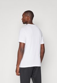 T-shirt bianco a maniche corte con scollo rotondo, caratterizzato da una texture liscia. Stile leggermente aderente, indossato con pantaloni scuri. Nessun motivo visibile.