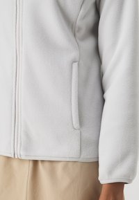 Giacca in fleece grigio chiaro con chiusura a zip, tasca laterale e rifiniture a contrasto sui polsini e sull'orlo. Texture morbida con una finitura liscia.