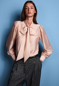 Gaiši rozā blūze ar lielu loku pie kakla, pūkainām piedurknēm un gludu tekstūru, kopā ar pelēkām augsta vidukļa biksēm ar ķēdes akcentu.