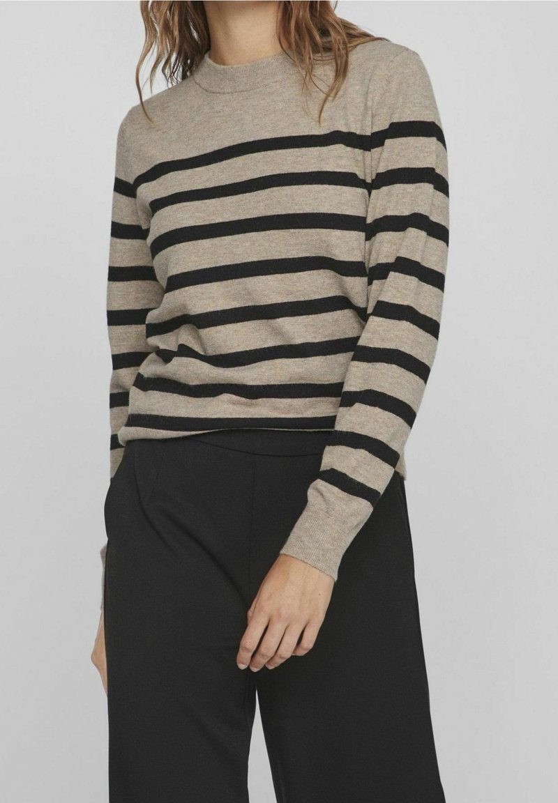 Personne portant un pull beige à manches longues avec des rayures horizontales noires et un pantalon noir, debout devant un fond uni.