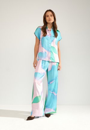 Femme portant une chemise ample à manches courtes boutonnée et un pantalon large avec un motif abstrait pastel bleu, rose et vert, debout sur un sol blanc.