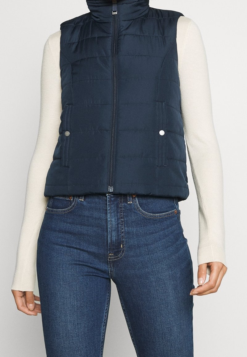 Donkerblauw gewatteerd vest met hoge kraag, ritssluiting en twee zakken. Heeft een horizontaal doorgestikt patroon en drukknopaccenten.