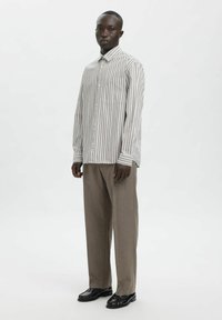 Moški stoji, oblečen v bež in belo črtasto srajco z gumbi, rjave ohlapne hlače in črne usnjene čevlje, na belem ozadju.