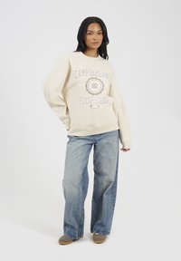 Donna che indossa una felpa color crema con la scritta "Camp Oakland Pennsylvania 1985", jeans a gamba larga bicolore blu e scarpe marroni, in piedi davanti a uno sfondo bianco.