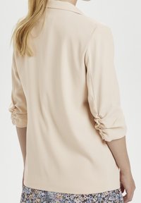 Žena s blond vlasy, oblečená v krémovém saku s nařasenými rukávy a modré květinové sukni, viděná ze zad.