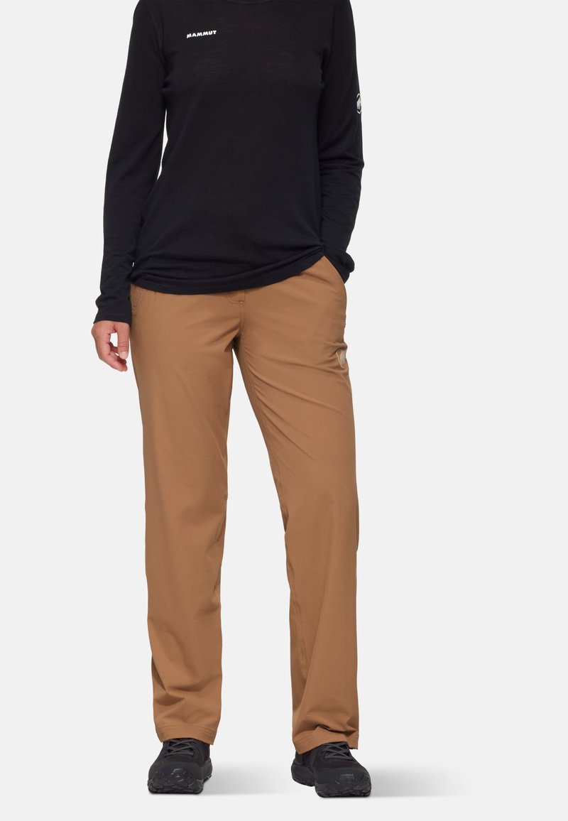 Personne portant un tee-shirt noir à manches longues et un pantalon beige droit avec des chaussures de randonnée noires, debout devant un fond uni.