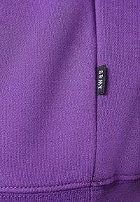 Tejido de punto púrpura con una etiqueta de ropa negra que dice "GRMY" unida a una costura.