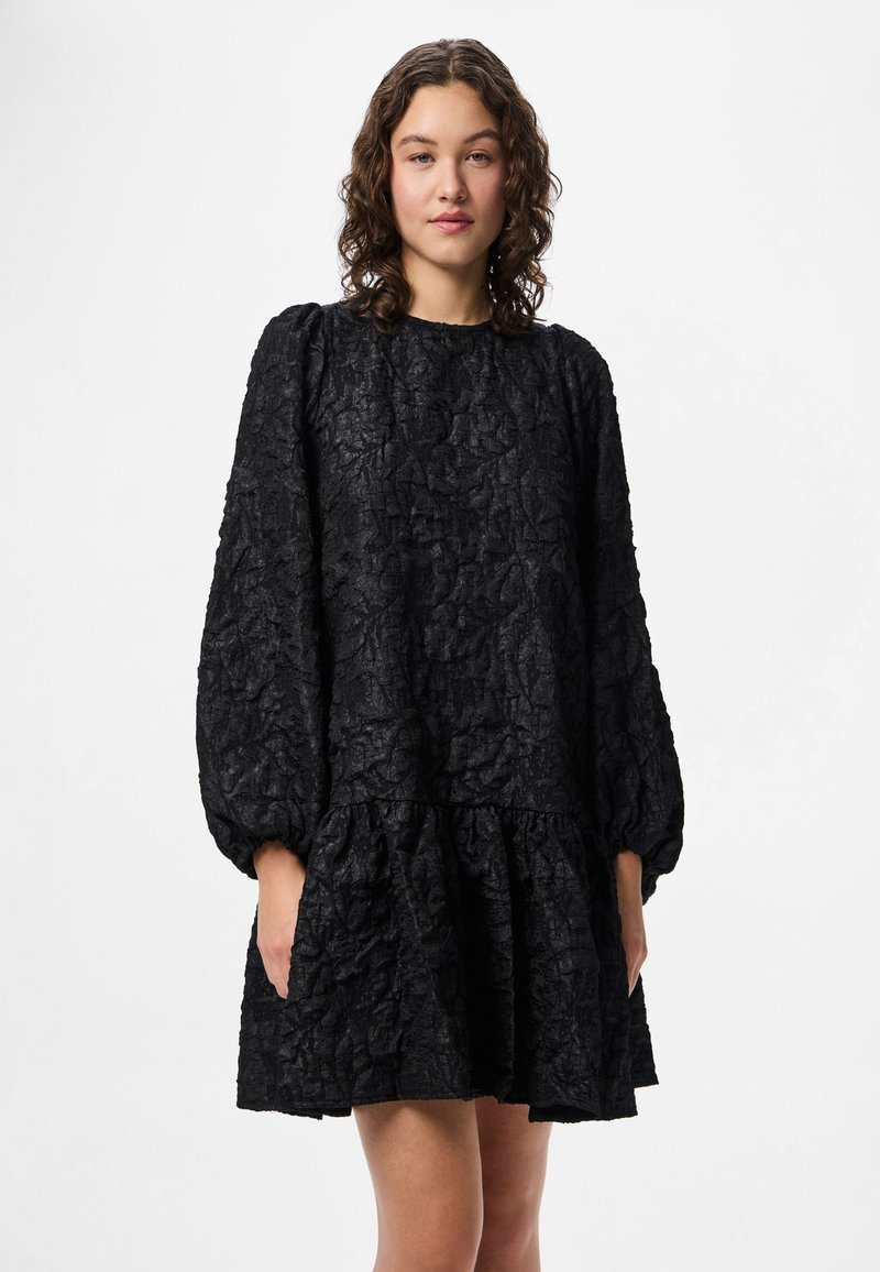 Crna teksturirana haljina s dugim balon rukavima, nabrane strukom i višeslojnim volanom na rubu. Izrađena od naborane tkanine, bez vidljivih kopčanja.