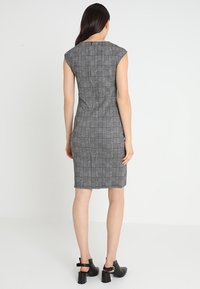 Robe ajustée à carreaux gris avec des manches courtes, longueur genoux et coutures sur les côtés. Portée avec des chaussures à talons blocs noires. Texture de tissu lisse.