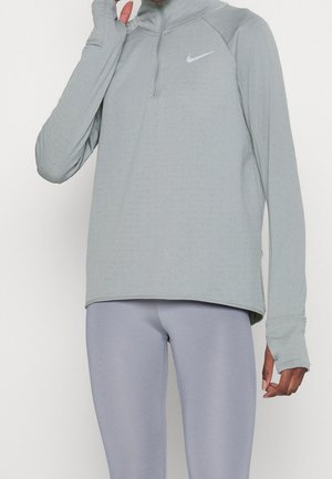 Personne portant un haut Nike à manches longues gris clair avec des trous pour les pouces et des leggings gris clair assortis, debout sur un fond blanc.