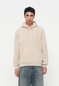 Beige Fleece-Hoodie mit Kängurutasche, Kapuze mit Kordelzug und gerippten Bündchen. Getragen mit hellblauen, lässig sitzenden Jeans.
