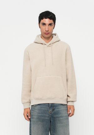 Beige fleece hoodie with a kangaroo pocket, drawstring hood, and ribbed cuffs. Worn with light blue loose-fitting jeans.
