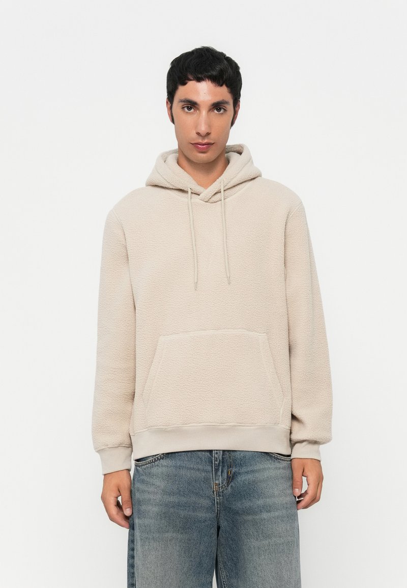 Beige Fleece-Hoodie mit Kängurutasche, Kapuze mit Kordelzug und gerippten Bündchen. Getragen mit hellblauen, lässig sitzenden Jeans.