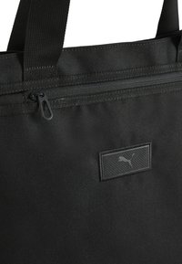 Sac tote noir fabriqué dans un tissu durable avec une finition texturée, doté d'une fermeture éclair supérieure et d'un patch logo rectangulaire.