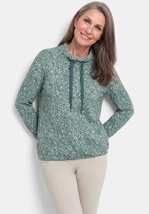 Lächelnde Frau mit langen grauen Haaren, die einen grünen gemusterten Pullover mit Kordeln und hellbeige Hose trägt, stehend mit den Händen hinter dem Rücken.