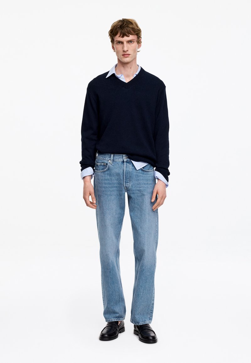 Ung man som bär en svart V-ringad tröja över en ljusblå skjorta, ljusblå jeans och svarta läderskor, stående mot en vit bakgrund.