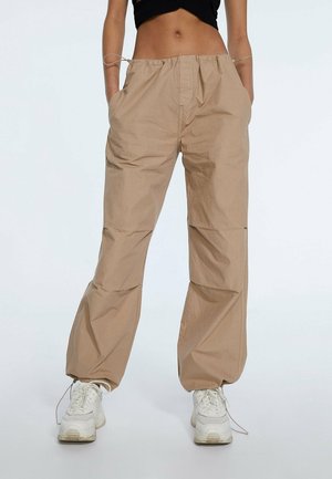Persona vistiendo pantalones beige sueltos con cintura elástica y zapatillas blancas, de pie con las manos en los bolsillos contra un fondo liso.