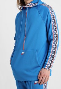 Sudadera con capucha azul que presenta un cierre de cremallera, bolsillo frontal y paneles laterales con diseño. Fabricada con tejido suave y detalles acanalados.